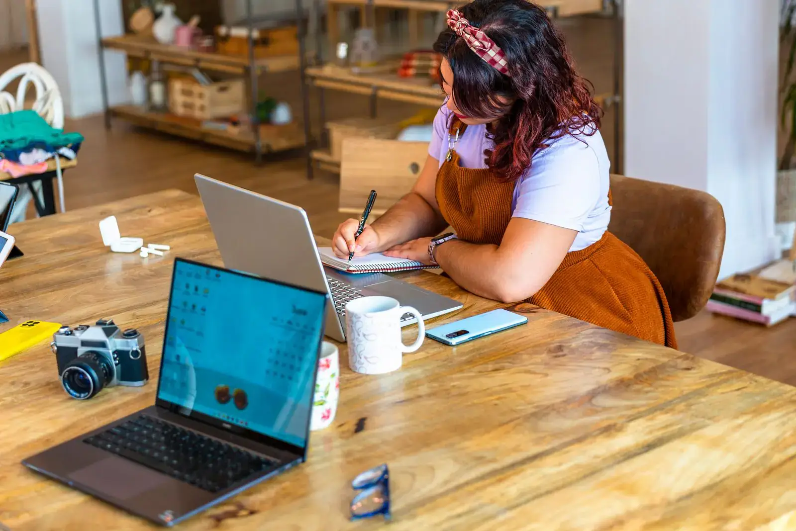 Frau schreibt Notizen an einem Holztisch mit zwei Laptops und einer Kamera, Symbolbild für den Blog eines kleinen Betriebs