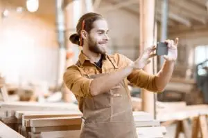 L&auml;chelnder Schreiner fotografiert in seiner Werkstatt mit dem Smartphone, Symbolbild f&uuml;r Social Media im Handwerk