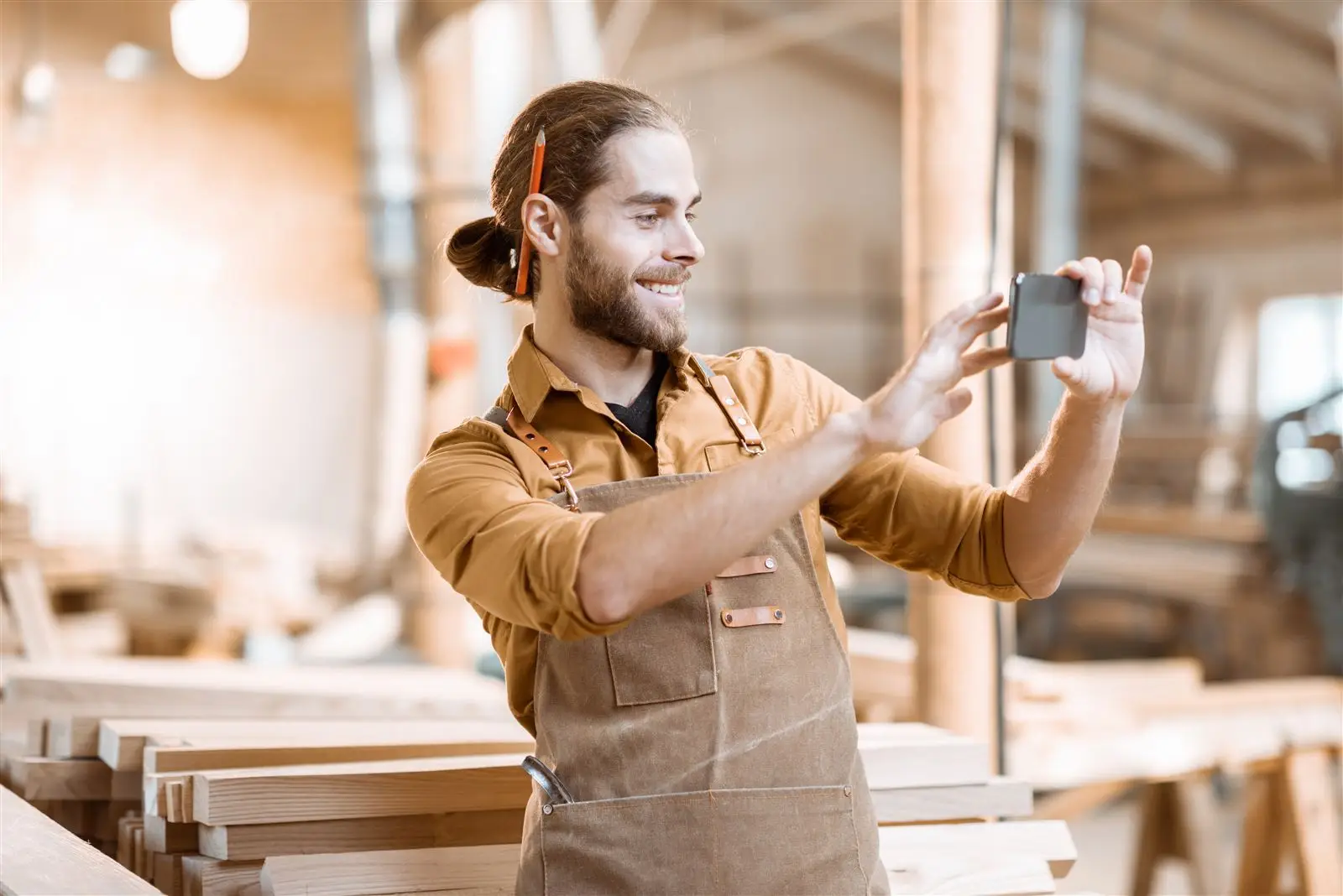Lächelnder Schreiner fotografiert in seiner Werkstatt mit dem Smartphone, Symbolbild für Social Media im Handwerk
